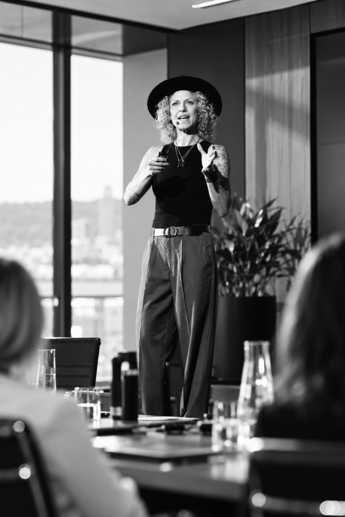 A confident speaker stands in front of an audience in a modern conference room, delivering a presentation. They have curly hair and wear a black hat, a sleeveless top, and loose trousers, with a backdrop of large windows showcasing a city view.